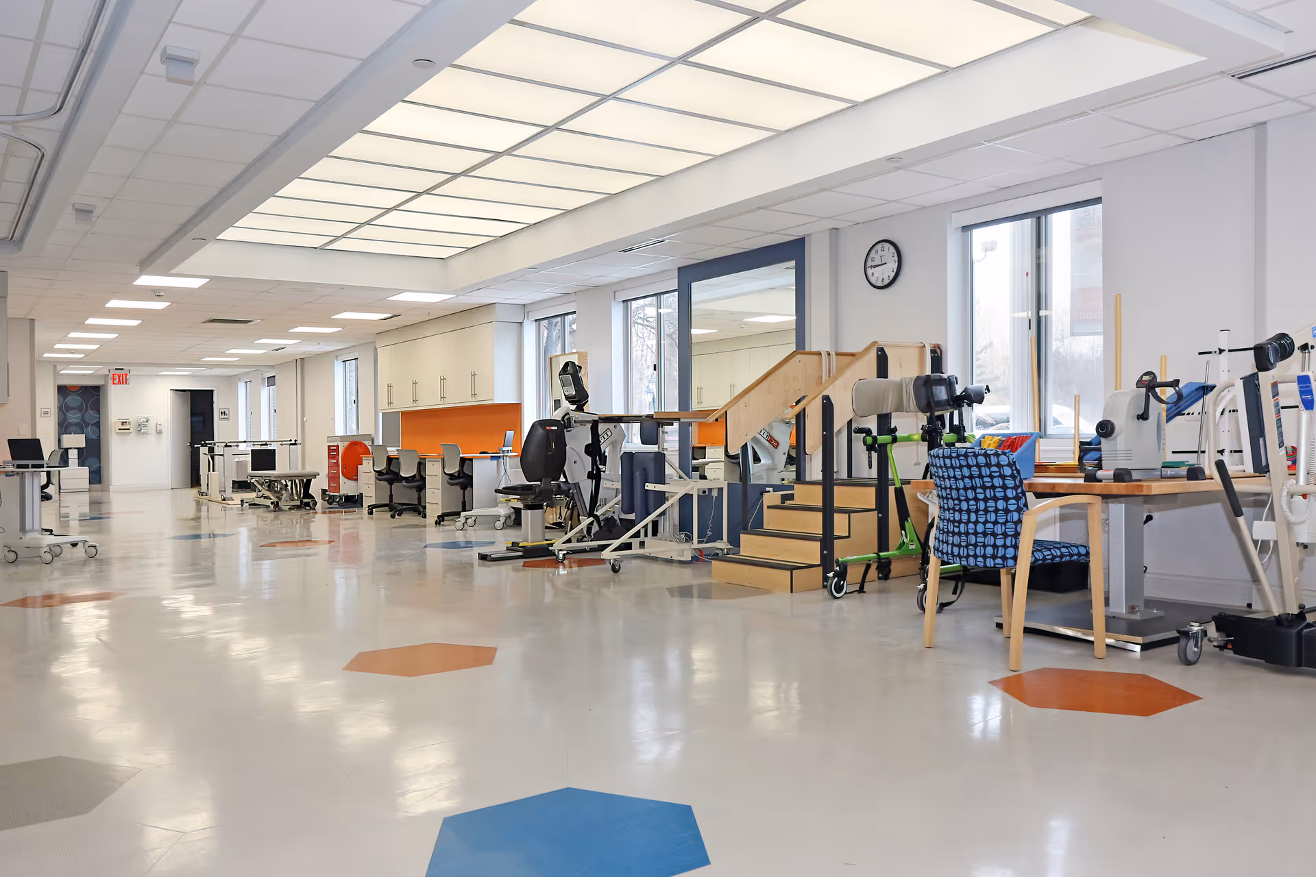 A spacious rehabilitation therapy room with exercise equipment, therapy stairs, chairs, desks, and large windows letting in natural light. The floor has colorful hexagonal patterns, and the ceiling features bright panel lighting.