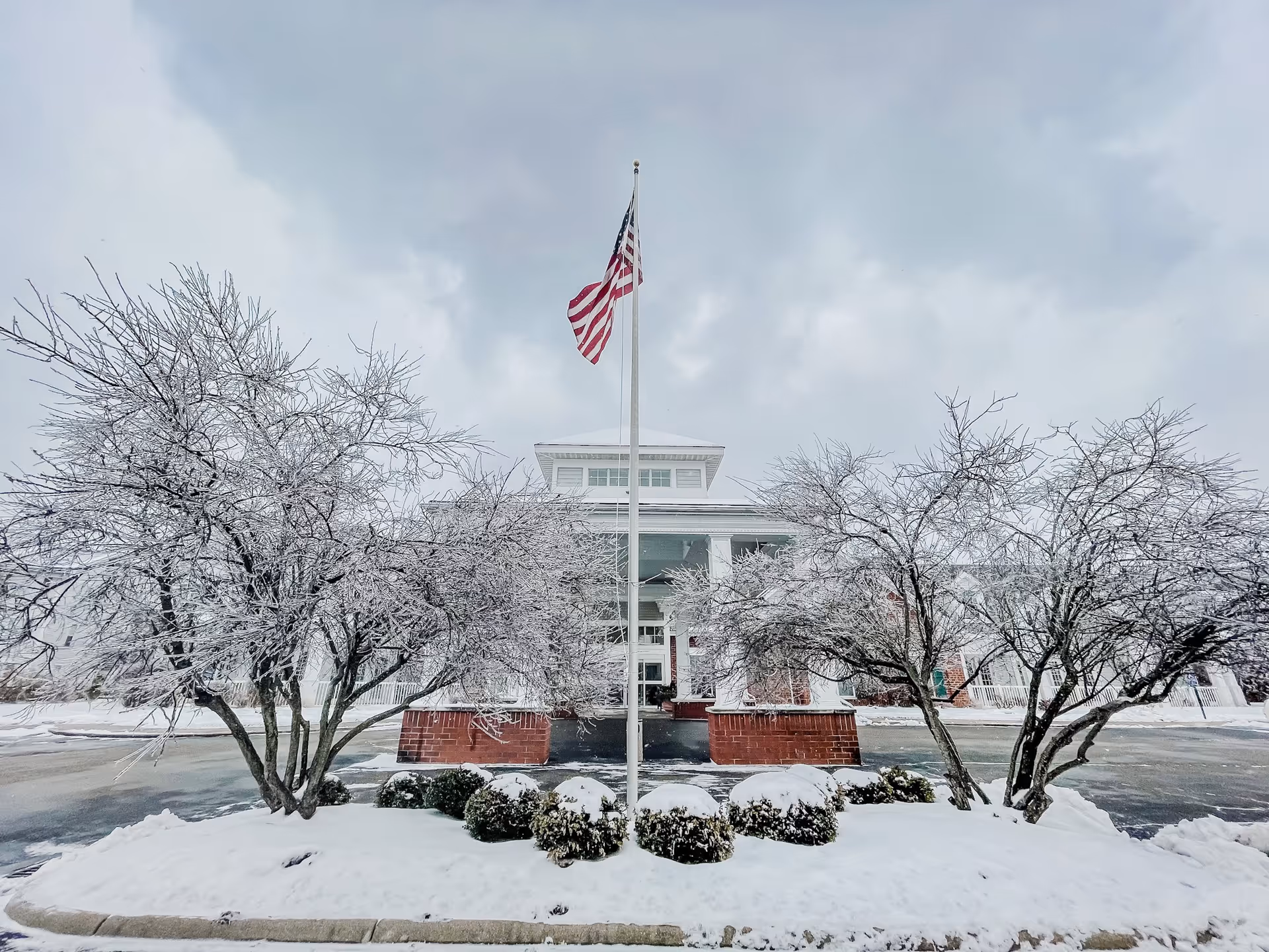 Snow-covered front entrance of a white community building with an American flag on a pole and frost-covered trees.