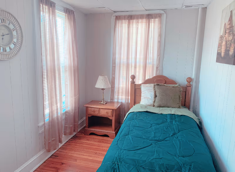 A small bedroom with a single wooden bed dressed in teal bedding, a wooden nightstand with a lamp, and two tall windows with sheer curtains.