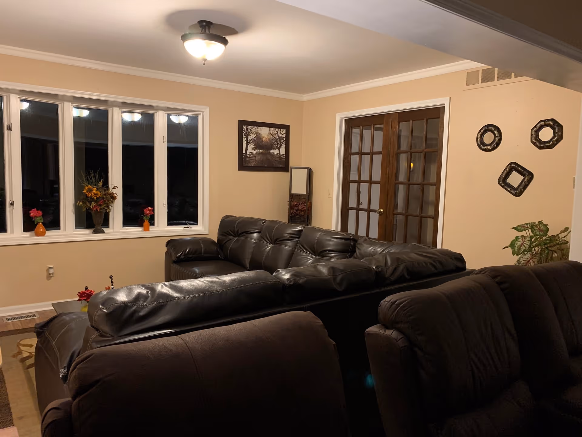 A cozy living room with beige walls, a ceiling light fixture, and a set of large windows with white trim. The room features dark leather sofas arranged around a coffee table with small flower vases. On one wall, there is a framed picture of a tree-lined path and a tall mirror with decorative items below it. French doors with glass panes lead to another room. Decorative wall hangings and a potted plant are also visible.