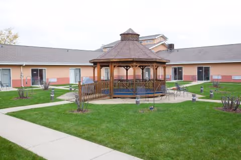 Outdoor courtyard area with a wooden gazebo in the center surrounded by green grass, paved walkways, and a single-story building with multiple doors and windows in the background.