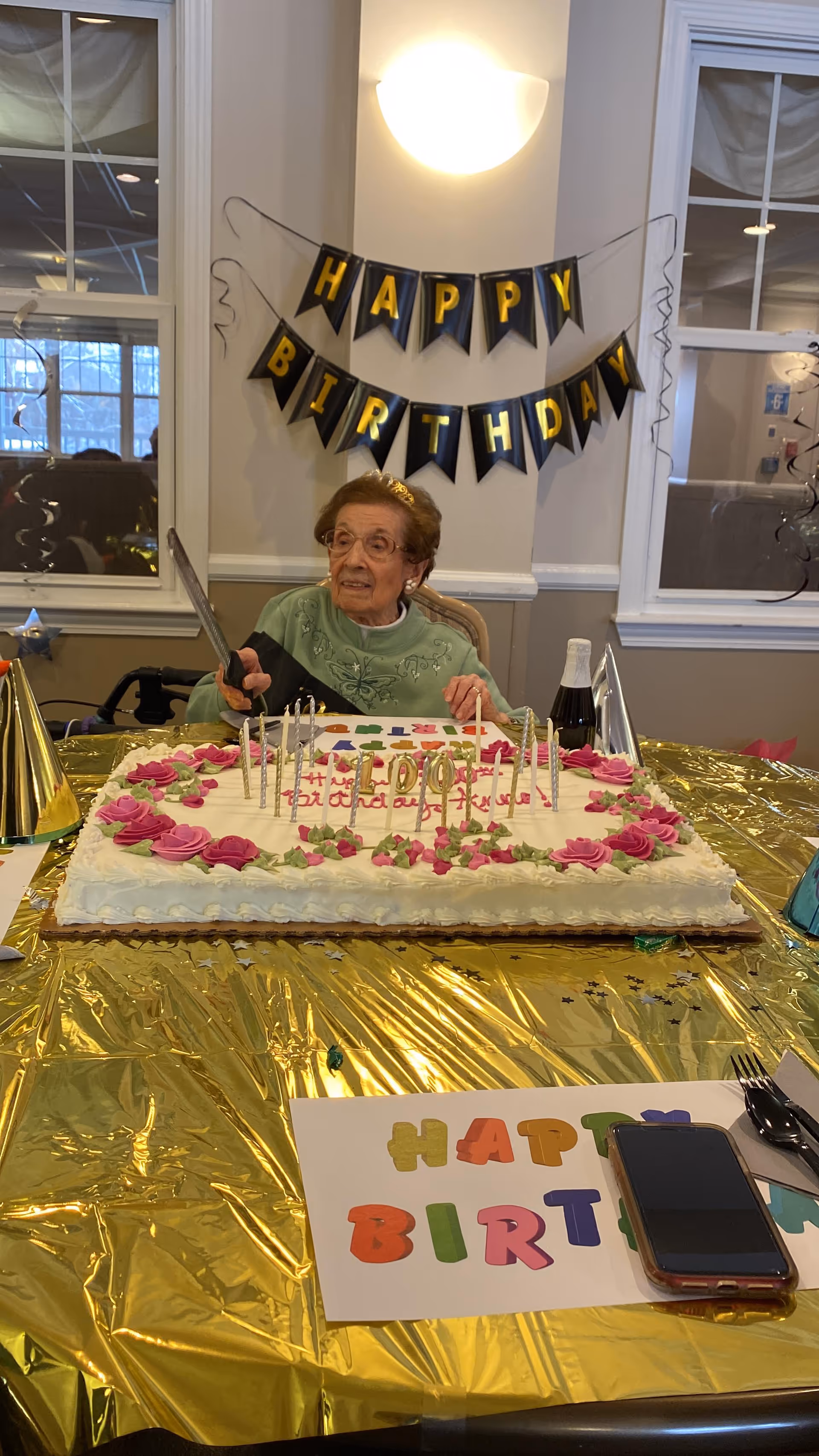 An elderly woman wearing a green sweater and a small tiara sits at a table with a large birthday cake decorated with pink roses and candles. Behind her, a black and gold banner reads 'HAPPY BIRTHDAY.' The table is covered with a shiny gold tablecloth and has party hats, a bottle, and a colorful 'HAPPY BIRTHDAY' sign along with a smartphone.