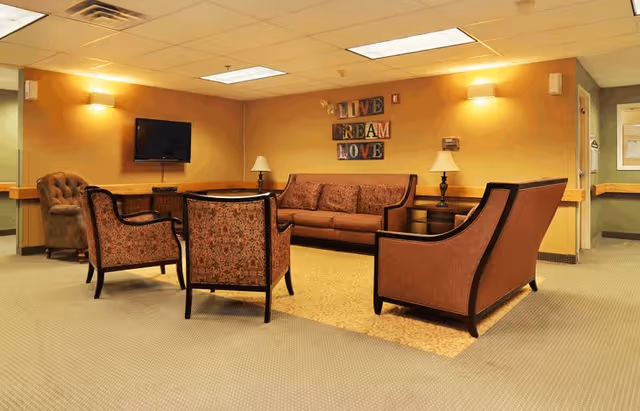 A cozy living room area with a brown sofa, three matching armchairs, two side tables with lamps, a wall-mounted TV, and a decorative wall sign that reads 'LIVE DREAM LOVE'. The walls are painted a warm yellow, and the floor is carpeted with a beige rug under the seating area.
