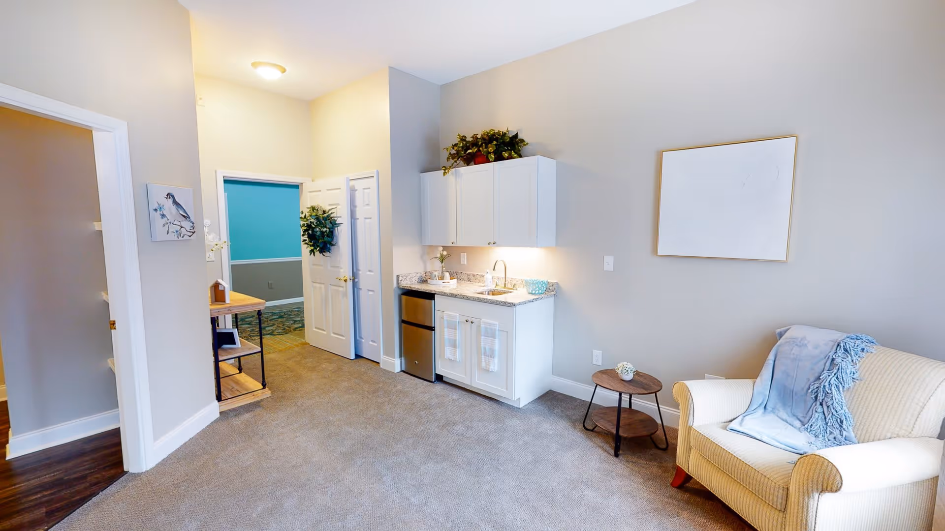A cozy interior room featuring a small kitchenette with white cabinets, a sink, and a mini refrigerator. To the right, there is a beige armchair with a blue throw blanket and a small round wooden side table. The walls are painted light gray, and there is a blank framed picture hanging above the armchair. The floor is carpeted, and there are two open doorways leading to other rooms, one with a blue wall visible. A small wooden table with shelves and decorative items is placed near one doorway.