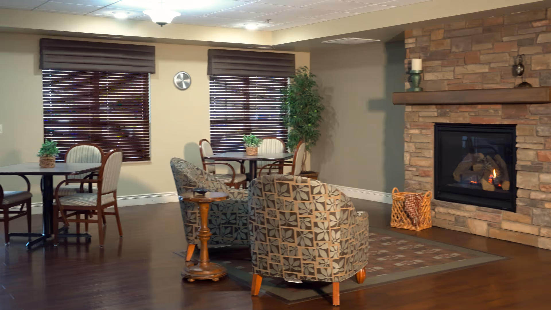 Cozy senior living lounge with patterned armchairs, small dining tables, and a stone fireplace.