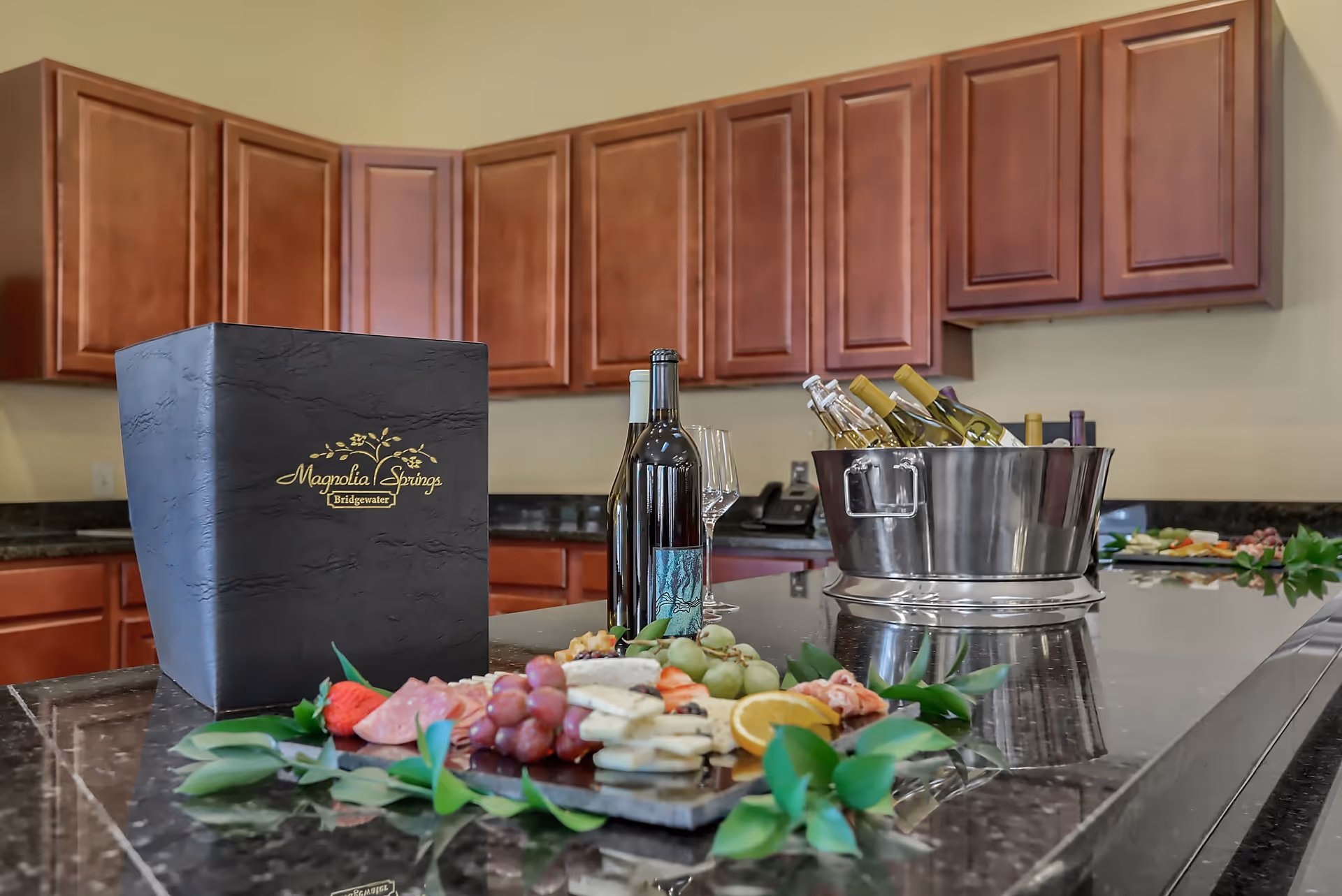 A kitchen countertop with a black box labeled Magnolia Springs Bridgewater, bottles of wine, wine glasses, a metal bucket filled with white wine bottles, and a charcuterie board with grapes, cheese, sliced meats, and orange slices. Wooden cabinets are visible in the background.