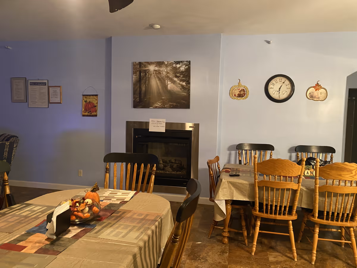Cozy dining area with two tables covered in tablecloths, wooden chairs, a fireplace and wall clock on a pale blue wall.