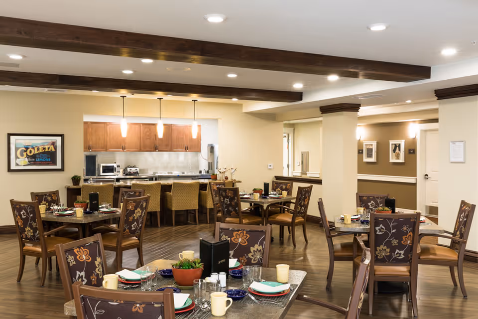 A well-lit dining room in a senior living facility with several tables set for meals. Each table has floral-patterned chairs, place settings with plates, cups, glasses, and napkins. In the background, there is a kitchen area with wooden cabinets, a microwave, and a toaster. The room features wooden beams on the ceiling and framed artwork on the walls.