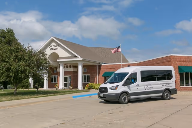 Brick senior living facility entrance labeled 'Colonial Manor' with an American flag and a white shuttle van parked in front.