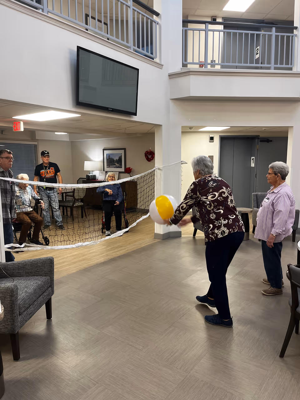 Seniors playing indoor beach-ball volleyball over a net in a community living area with seating and a wall-mounted TV.