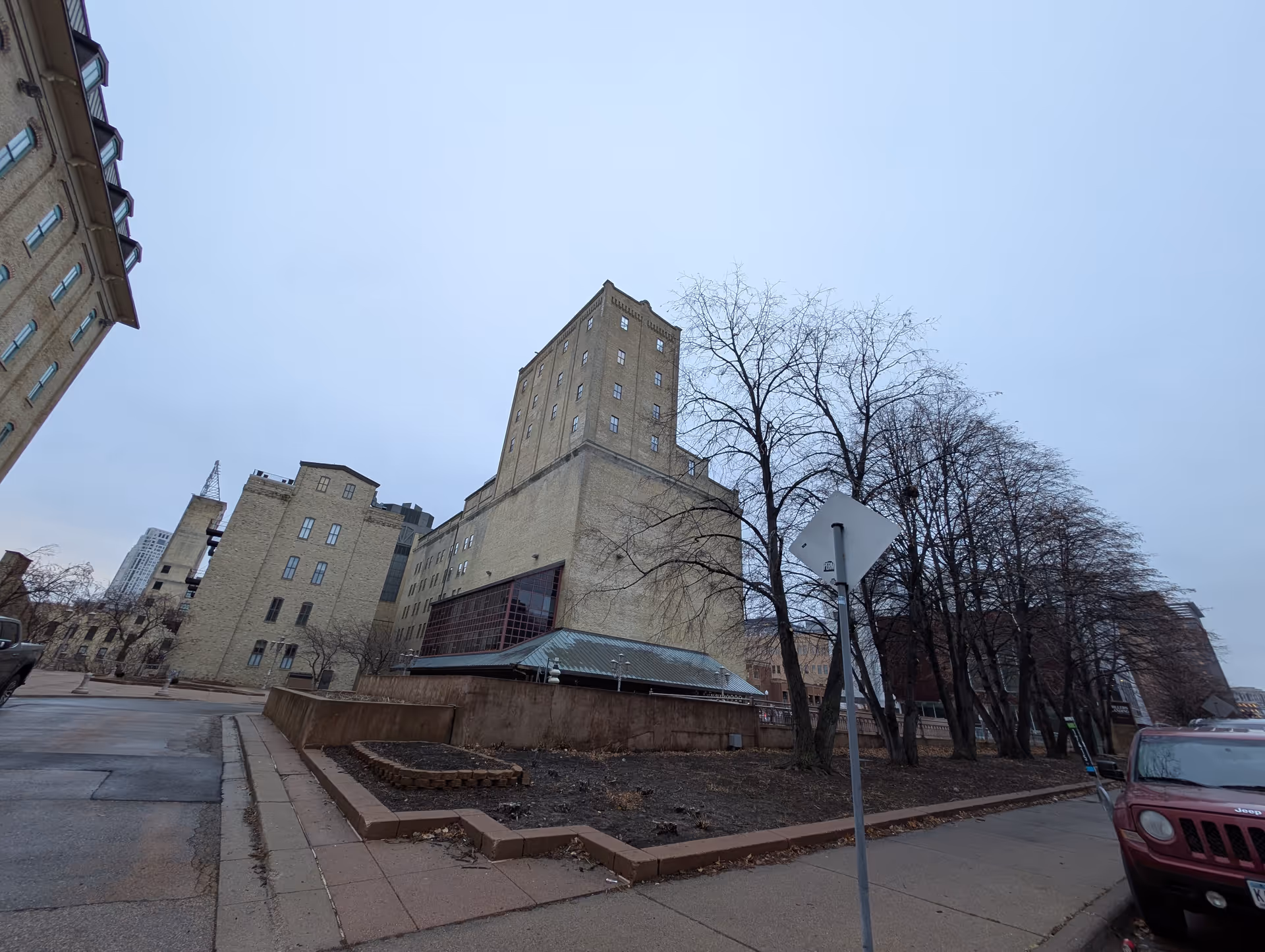 Exterior view of a large multi-story brick building with several windows, adjacent to a sidewalk lined with leafless trees and a parked red Jeep. The sky is overcast.