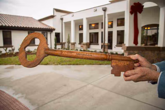 A person holding a large wooden ceremonial key in front of a white building with a tiled roof and columns, likely the entrance to a senior living facility.