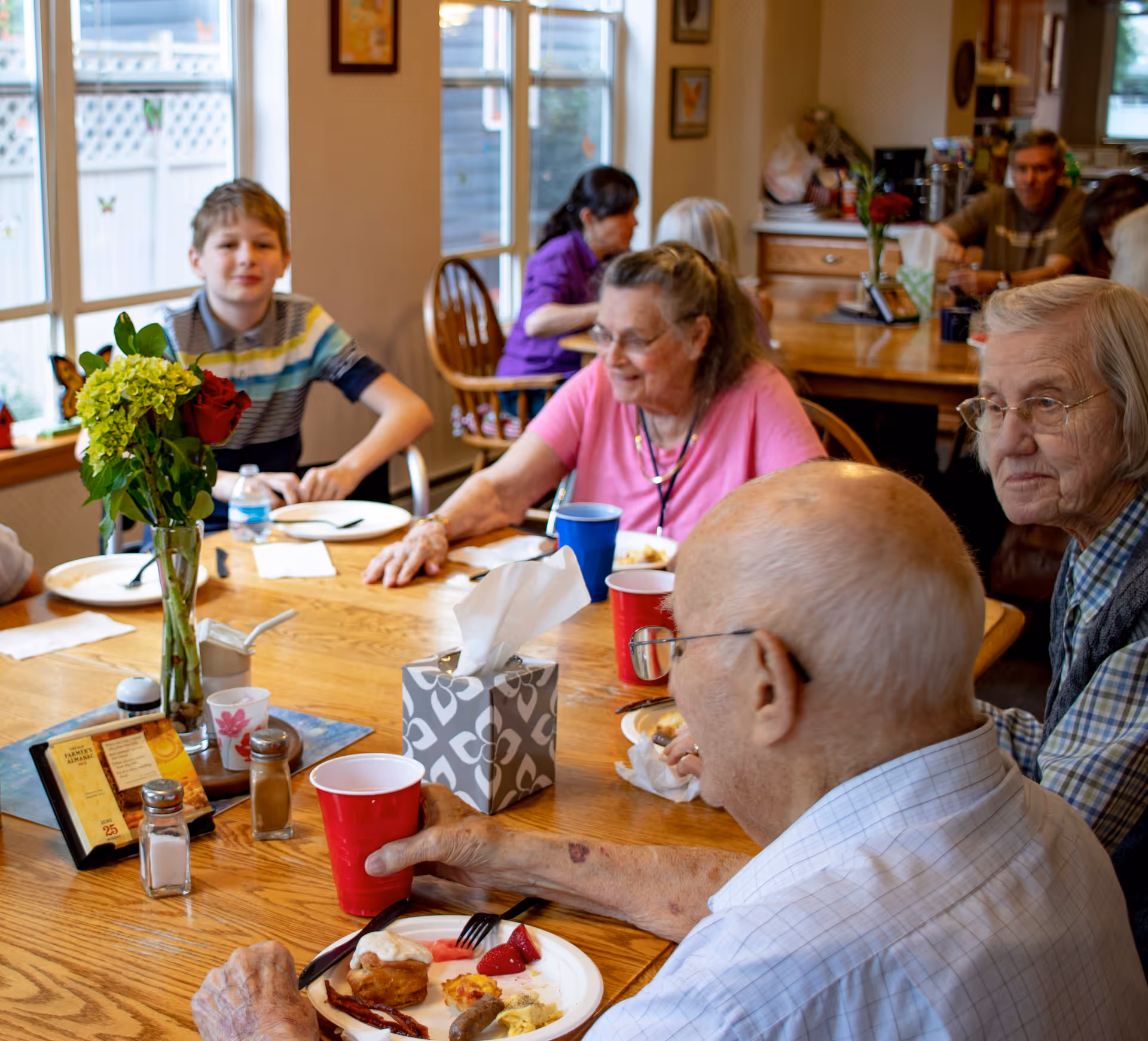 A group of elderly people and a young boy sitting around a wooden dining table enjoying a meal together in a well-lit room with large windows. The table has plates with food, cups, a tissue box, salt and pepper shakers, and a vase with flowers. The atmosphere appears warm and social.
