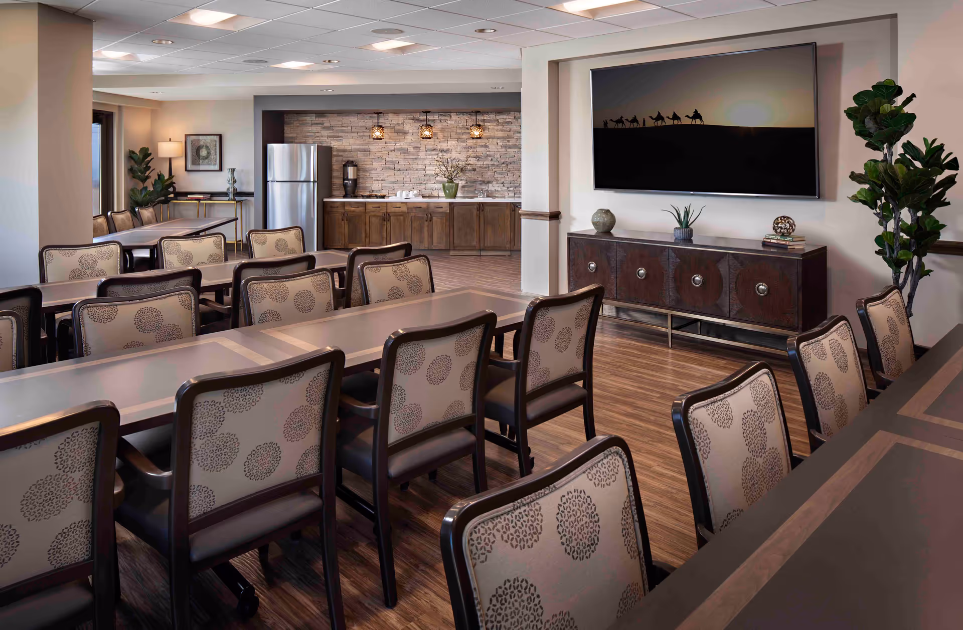 A spacious dining area in a senior living facility with multiple tables and chairs arranged neatly. In the background, there is a kitchenette with a refrigerator, coffee maker, and wooden cabinets under a stone accent wall. On the right side, a large flat-screen TV is mounted on the wall above a decorative wooden console with plants and books. The room has wood flooring and soft lighting.