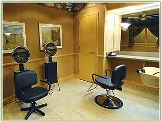 Interior view of a salon area with two black salon chairs, two hair dryer stations, a large mirror, and a sink for hair washing. The walls are painted beige with framed pictures and a door in the background.