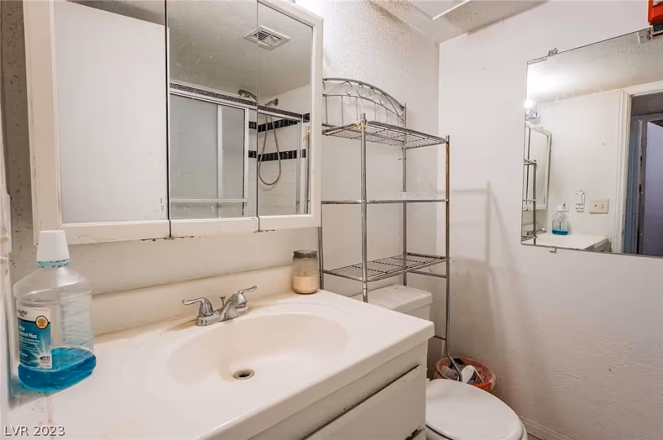 A small bathroom with a white sink and countertop, a silver faucet, a mirrored medicine cabinet above the sink, a metal shelving unit over the toilet, and a shower with sliding glass doors in the background.