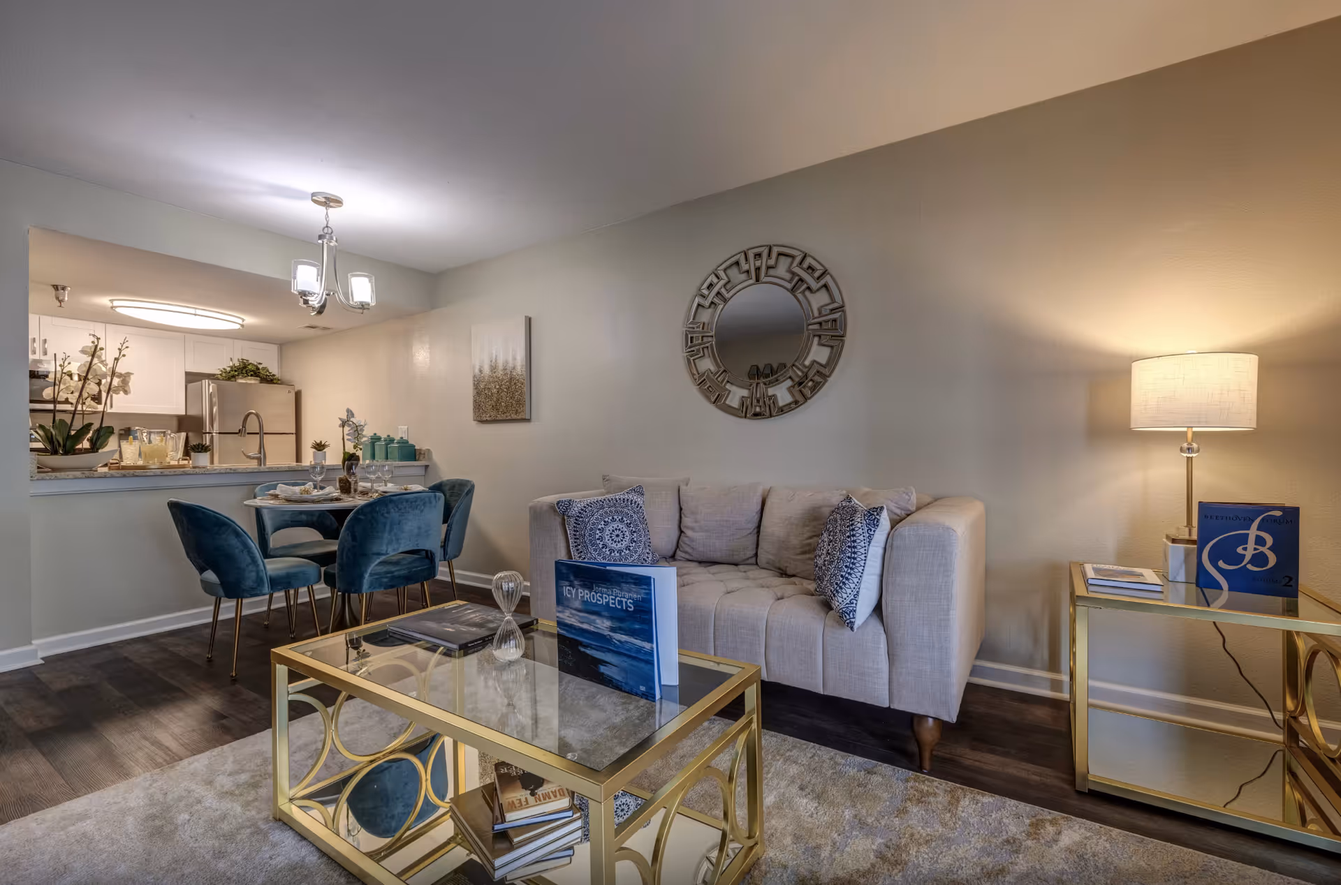 Open-concept living room and dining area with a beige sofa, gold coffee table, blue dining chairs and a view into the kitchen.
