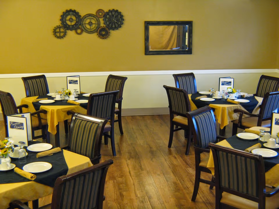 Dining room with several tables covered with yellow and black tablecloths, set with plates, cups, napkins, and small flower arrangements. The room has wooden flooring, beige walls with a white chair rail, a decorative wall clock made of gears, and a rectangular mirror.