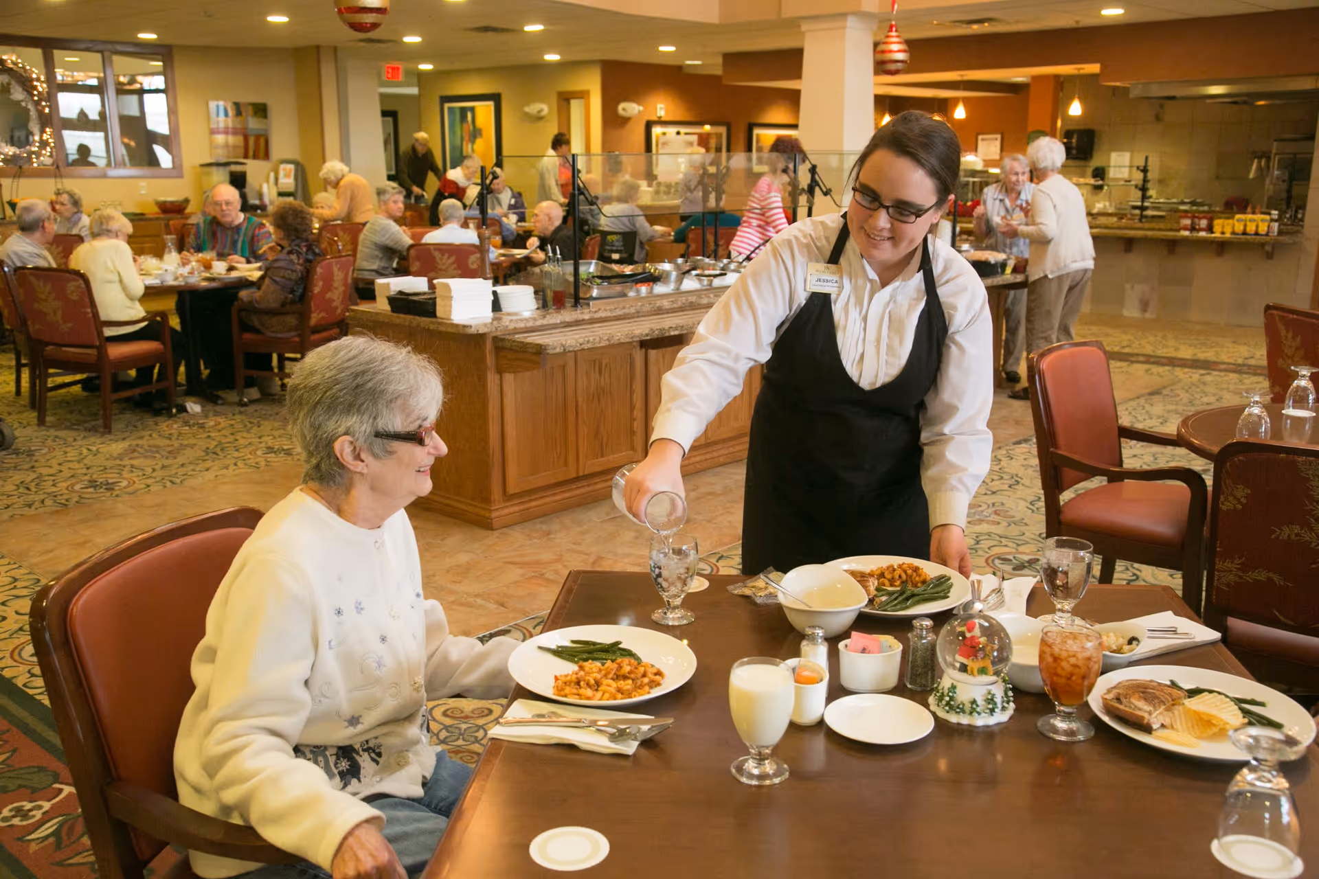 A dining room in a retirement community where an elderly woman is seated at a table with plates of food. A staff member wearing a black apron and white shirt is pouring water into a glass for the woman. Other elderly residents are seated at tables in the background, eating and socializing.