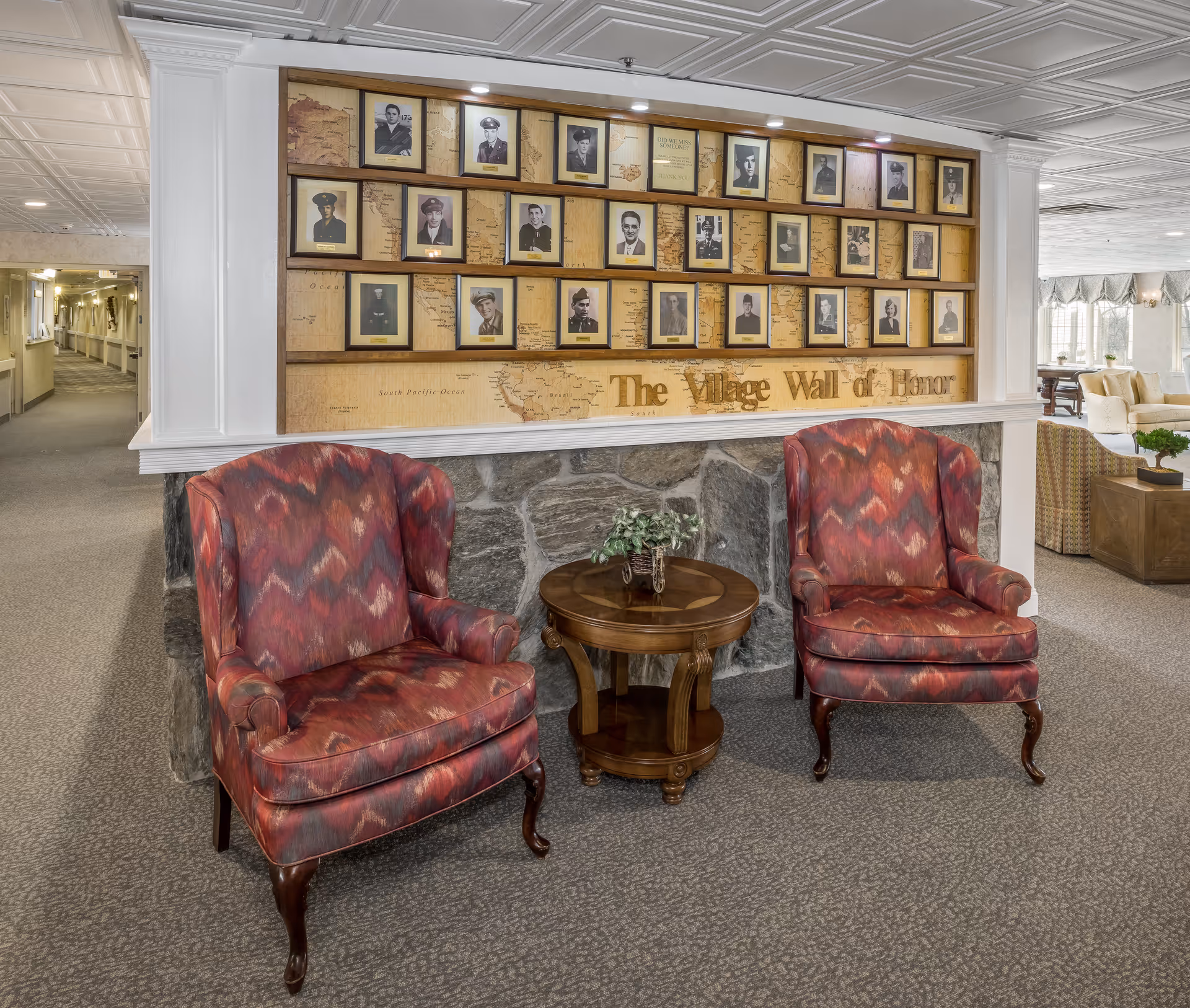 Two patterned armchairs with wooden legs are positioned on either side of a small round wooden table with a plant on it. Behind the chairs is a stone half-wall topped with a display titled 'The Village Wall of Honor' featuring framed black and white portraits of veterans. The setting appears to be a cozy interior common area with carpeted floors and soft lighting.
