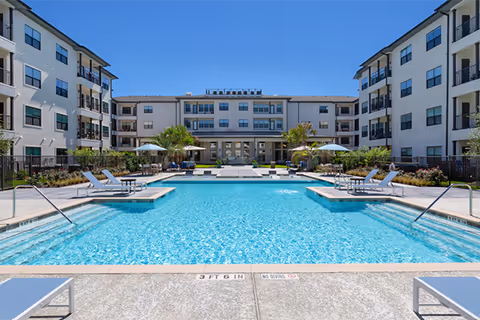 Large outdoor swimming pool surrounded by lounge chairs and umbrellas in the courtyard of a multi-story senior living building.