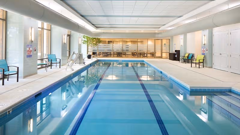 Indoor swimming pool with clear blue water, surrounded by a tiled deck with chairs along the sides. Large windows on the left side allow natural light to enter. A glass wall at the far end separates the pool area from a room with tables and chairs. The ceiling has large white panels with lighting.