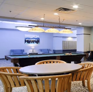 A recreational room with a pool table in the center, overhead lighting with four lamps, a round table with wooden chairs in the foreground, and a seating area with blue chairs and a lamp against a light purple wall with a landscape painting.