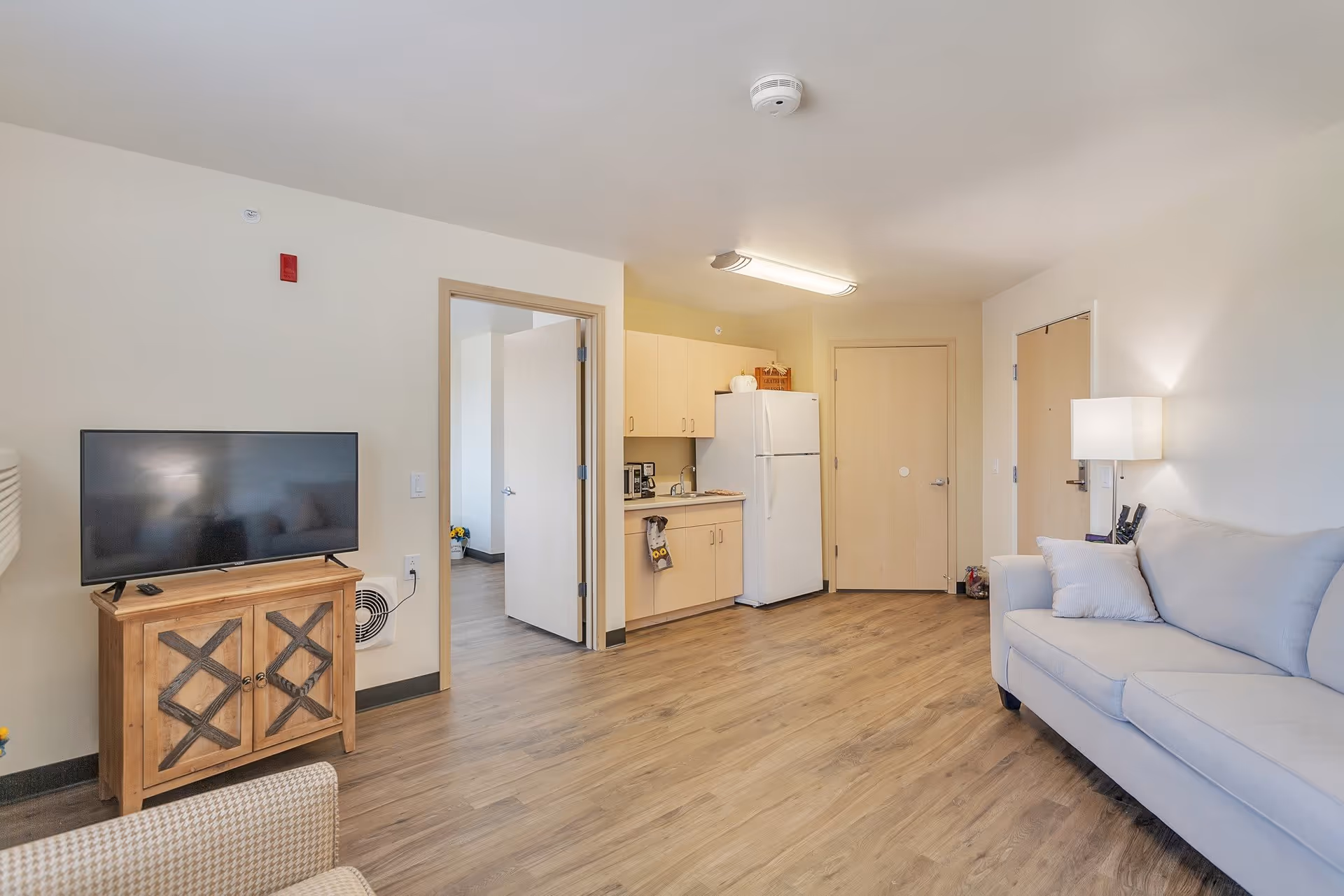 A bright and clean living area in a senior living facility featuring a light gray sofa with pillows on the right, a wooden TV stand with a flat-screen television on the left, and a small kitchenette with light wood cabinets, a white refrigerator, and a microwave in the background. The floor is wood laminate, and there is a doorway leading to another room.