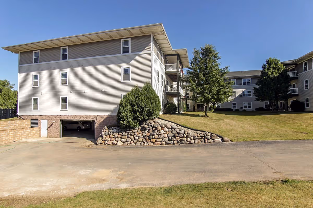 Three-story beige senior apartment building with balconies, a stone retaining wall, lawn, and trees under a clear blue sky.