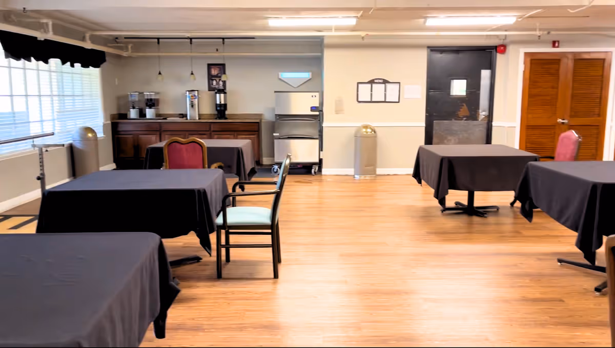 A dining room with several tables covered in black tablecloths and a mix of chairs around them. The room has wooden flooring, large windows with blinds on the left side, and a counter area at the back with coffee and beverage machines. There is a black door and wooden double doors on the back wall.