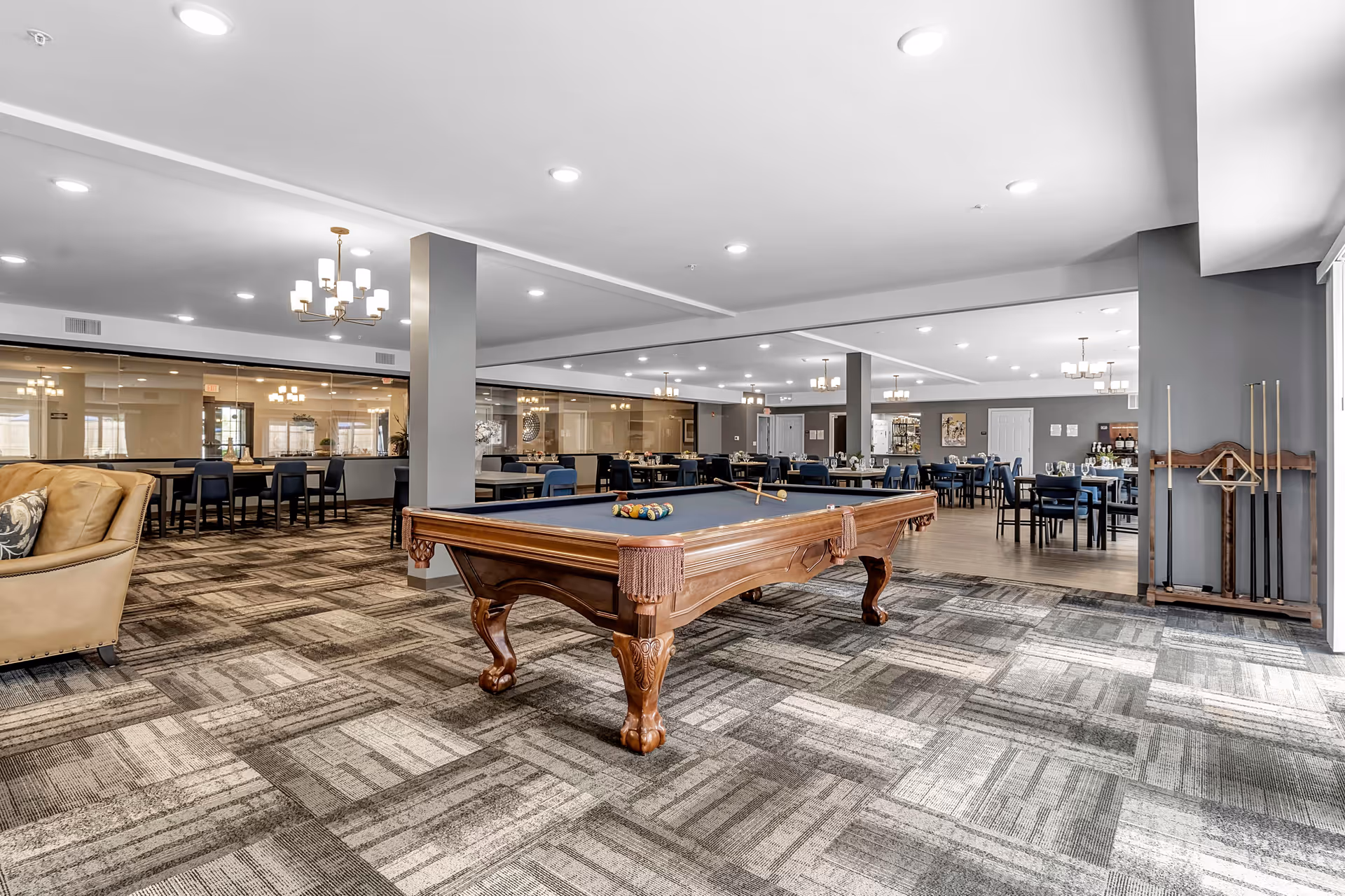 A spacious common area featuring a wooden pool table with blue felt in the center, surrounded by a patterned carpet. To the right, there is a rack holding pool cues. In the background, there are multiple dining tables and chairs arranged neatly under ceiling lights, with a large mirror reflecting the room. A beige armchair is partially visible on the left side.