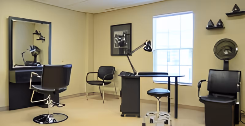 Interior view of a salon or grooming area with a black styling chair in front of a mirror, two additional black chairs, a small table with a desk lamp, and a window letting in natural light. The walls are beige with some decorative items mounted.