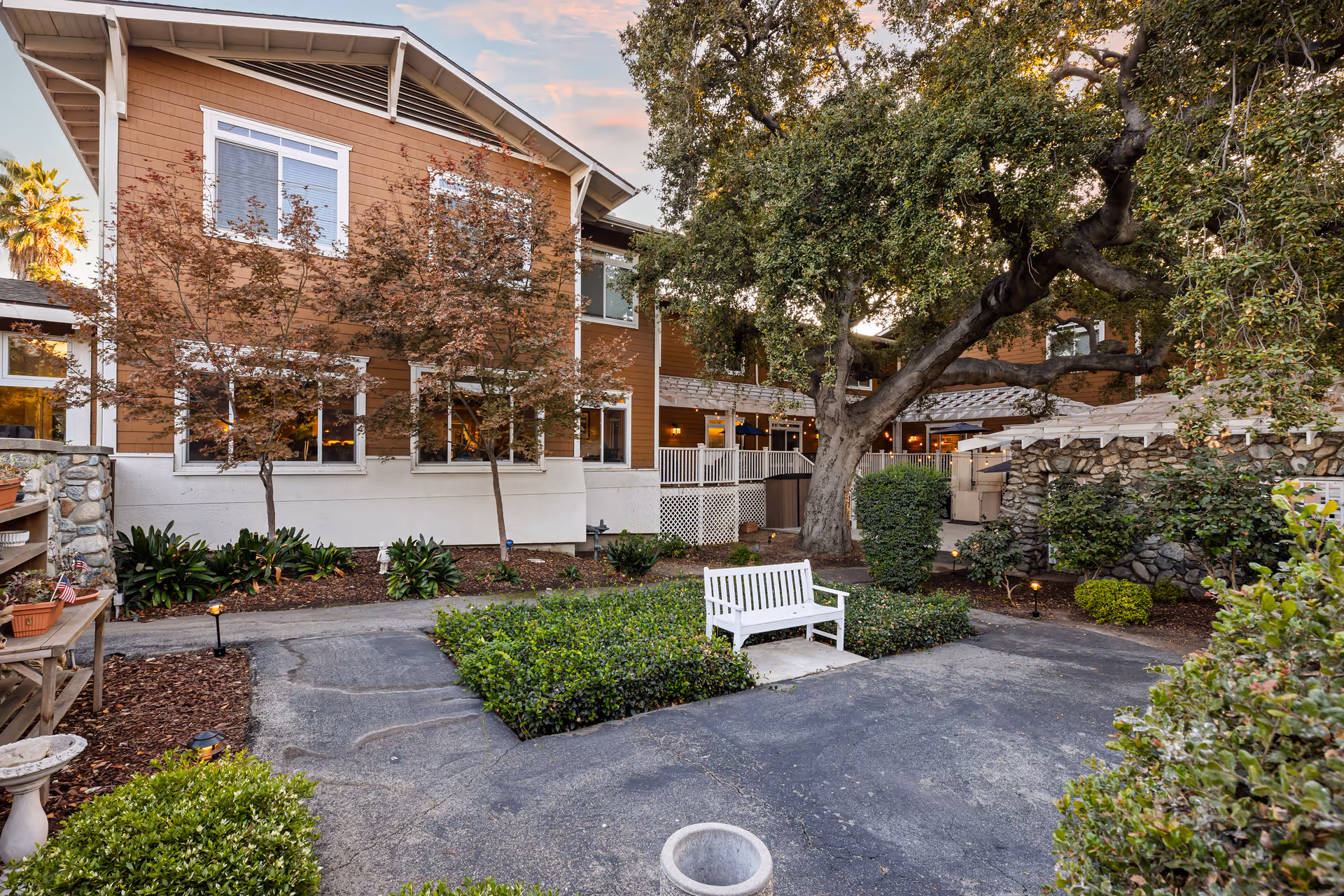 Outdoor courtyard area at Ivy Park at Claremont featuring a large tree, a white bench, various bushes and plants, and a two-story building with brown siding and white trim in the background under a partly cloudy sky.