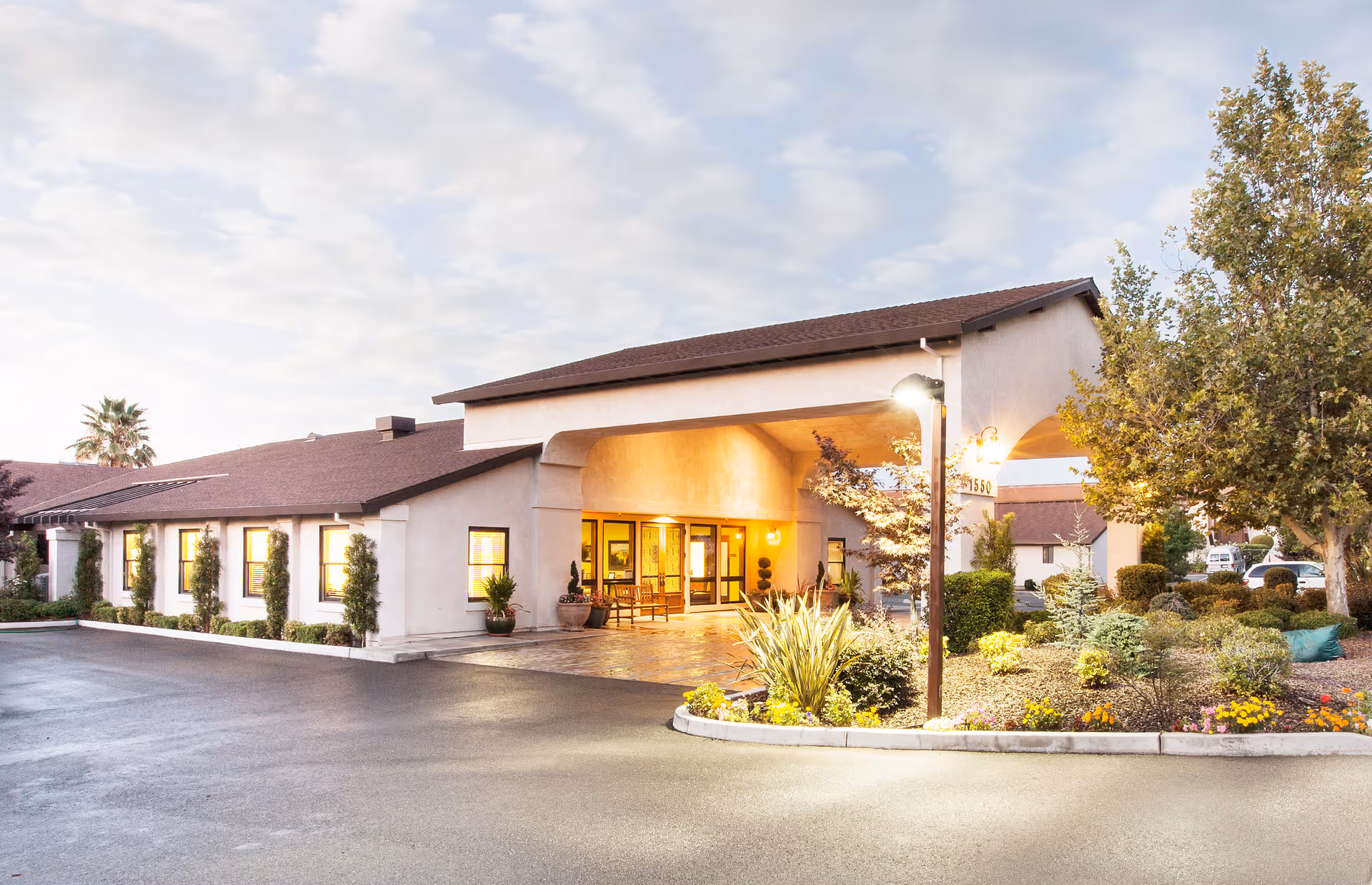 Exterior view of Lincoln Meadows Care Center building at dusk with warm lights glowing from inside. The building has a covered entrance with a tiled floor, surrounded by landscaped bushes, trees, and flowers. The sky is partly cloudy and the parking area is visible in front.