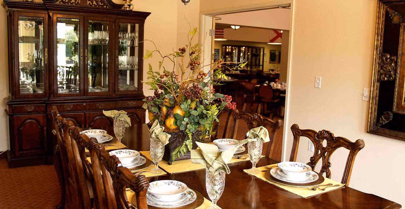 A formal dining room with a long wooden table set for six people with plates, bowls, glasses, and folded napkins. A large floral centerpiece with greenery and fruit decorates the middle of the table. In the background, there is a wooden china cabinet with glass doors displaying glassware. The room has warm lighting and a doorway leading to another dining area.