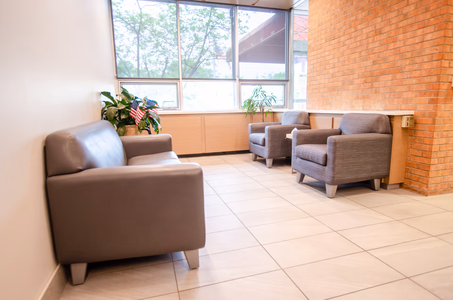 A bright waiting area with a gray leather sofa on the left and two gray upholstered armchairs on the right. There are large windows letting in natural light, a brick wall on the right, and potted plants including one with small American flags near the sofa.