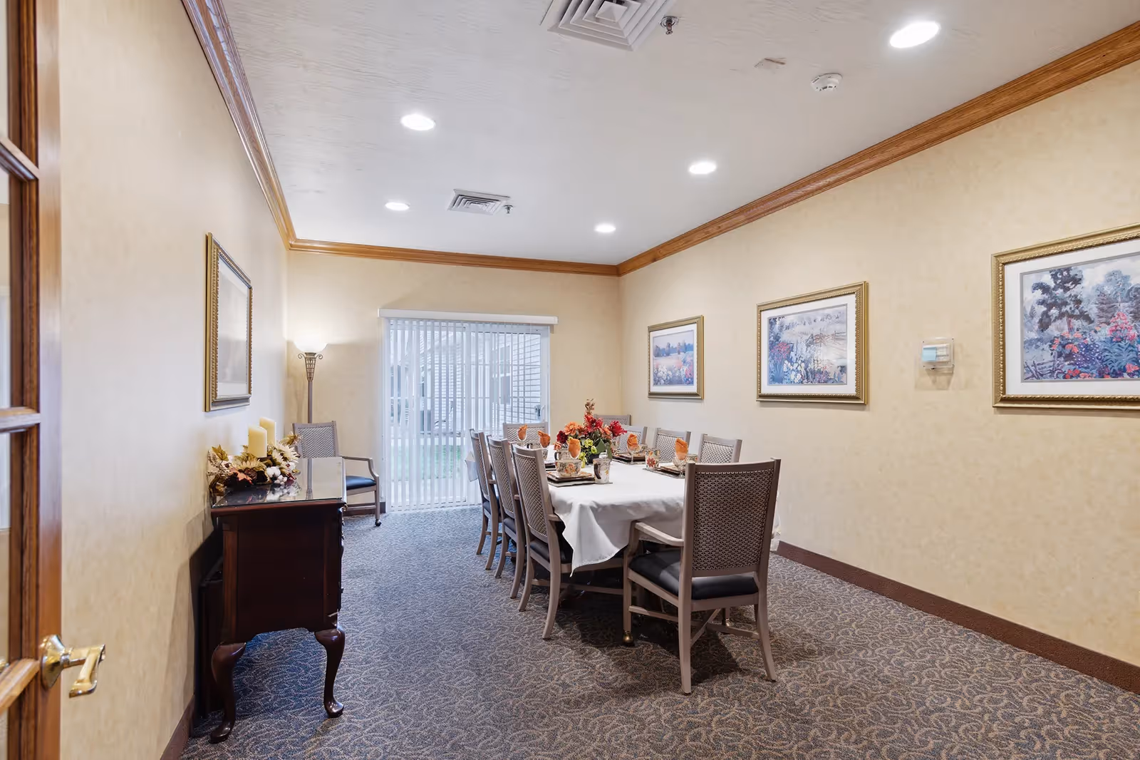 A dining room with a long rectangular table set for a meal, surrounded by chairs. The room has beige walls with framed artwork, a carpeted floor, recessed ceiling lights, and a sliding glass door with vertical blinds leading outside. A small wooden side table with decorative candles and flowers is positioned near the door.