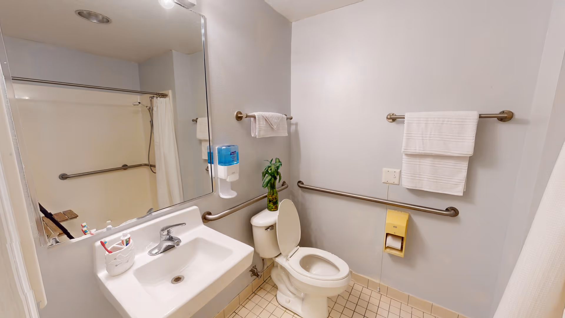 A clean bathroom with a white sink, a large mirror above it, a toilet with the lid open, grab bars on the walls, a towel rack with white towels, a soap dispenser mounted on the wall, and a shower area with a curtain in the background.