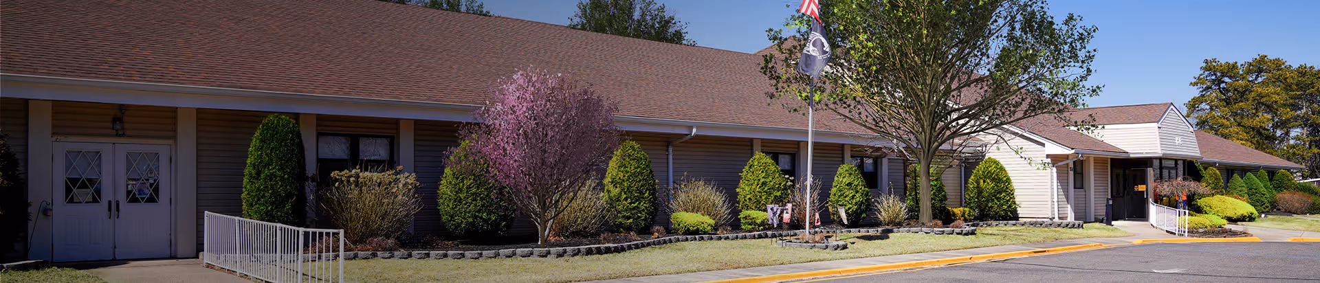Long, single-story senior living facility exterior with landscaped shrubs, a flagpole, and a walkway along the front.