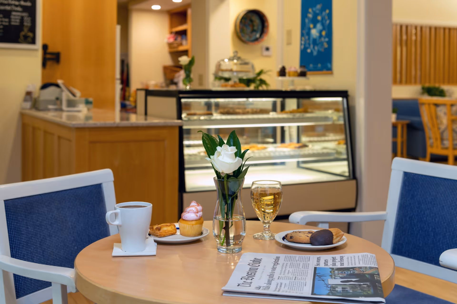A cozy cafe or dining area with a round wooden table set with a white coffee cup, a plate with a pastry and a cupcake, a glass of light-colored beverage, a plate with cookies and a pastry, a vase with a white rose, and a newspaper. In the background, there is a display case with baked goods and wooden furniture with blue cushions.