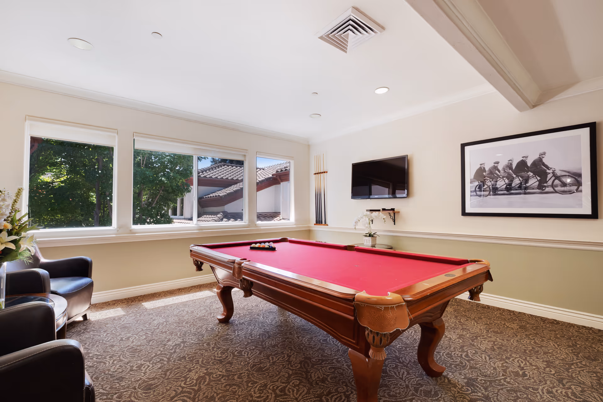 A well-lit game room with a red felt pool table in the center, two black leather chairs with a small round table between them, large windows showing trees and a tiled roof outside, a wall-mounted TV, a set of pool cues on the wall, and a black and white framed photo of five men riding a tandem bicycle.