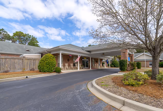 Exterior view of The Legacy of North Myrtle Beach senior living facility entrance with a covered drop-off area, rocking chairs on the porch, an American flag, and landscaped surroundings including bushes and a tree.