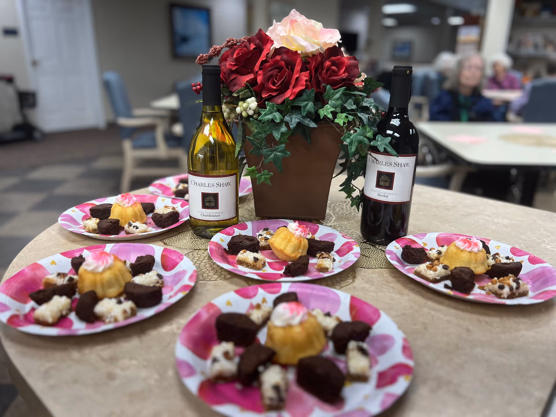 A round table with five pink and white patterned plates, each containing a small cake with pink frosting, brownies, and other dessert pieces. In the center of the table is a brown vase with red and white artificial flowers and green leaves. Two bottles of Charles Shaw wine, one white and one red, are placed on the table. In the background, there are blurred images of people sitting at tables in a communal dining or lounge area.