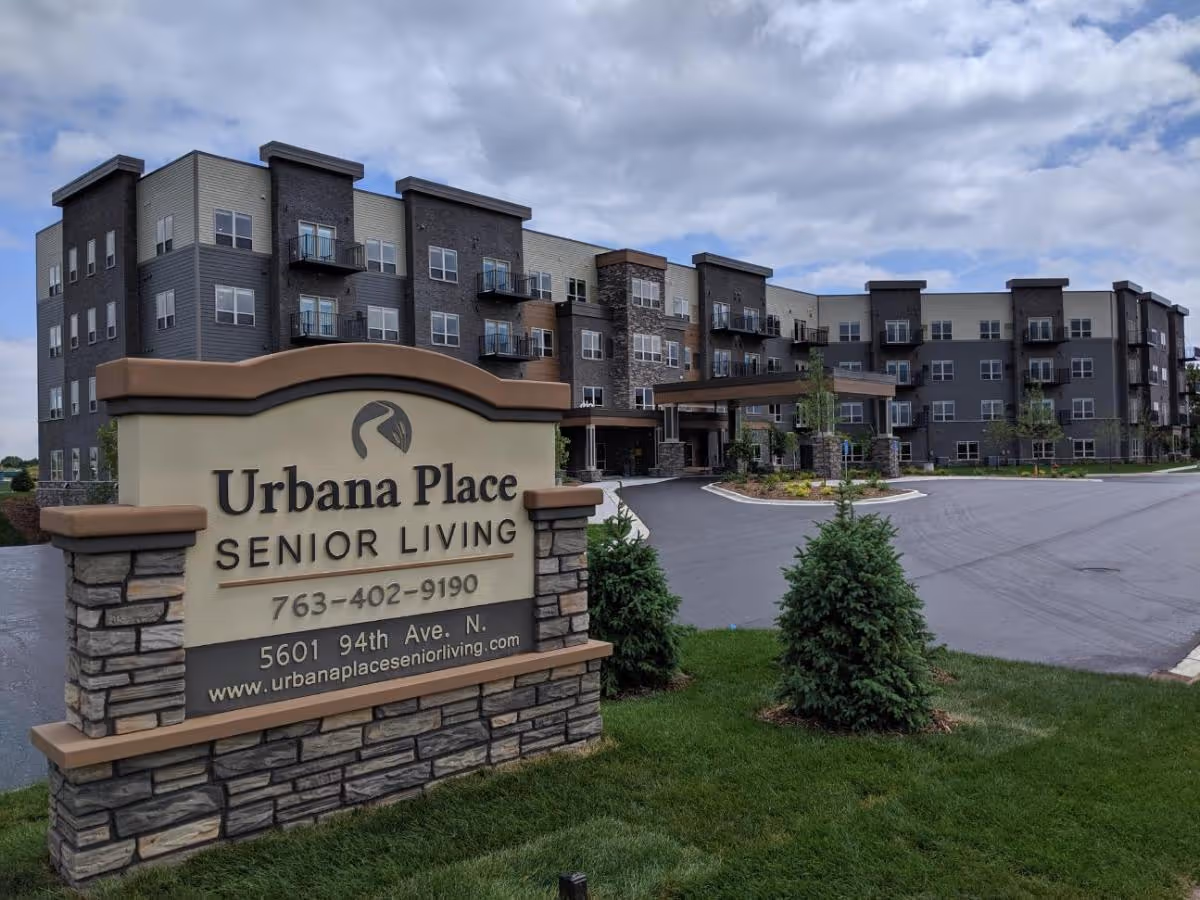 Exterior view of Urbana Place Senior Living building with a large sign in the foreground displaying the facility's name, phone number, address, and website. The building is multi-story with balconies and a covered entrance, surrounded by a paved driveway and landscaped greenery under a partly cloudy sky.