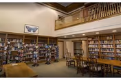 Interior view of a spacious library room with tall bookshelves filled with books along the walls. There is a wooden table with chairs in the center and a balcony railing above. A framed picture of a butterfly is hung on the wall above the bookshelves.