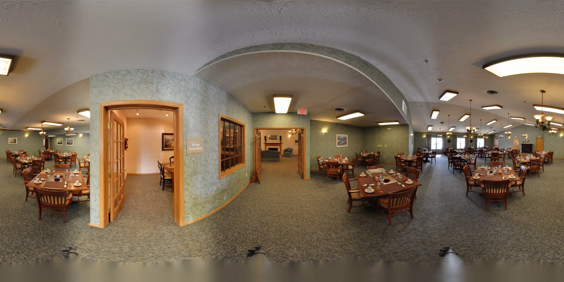 A spacious dining room in a senior living facility with multiple wooden tables and chairs arranged neatly. Each table is set with cups, saucers, and utensils. The room has soft green walls, carpeted floors, and ceiling lights. There are two doorways visible, one leading to a private dining room and another to a lounge area with sofas and a fireplace.