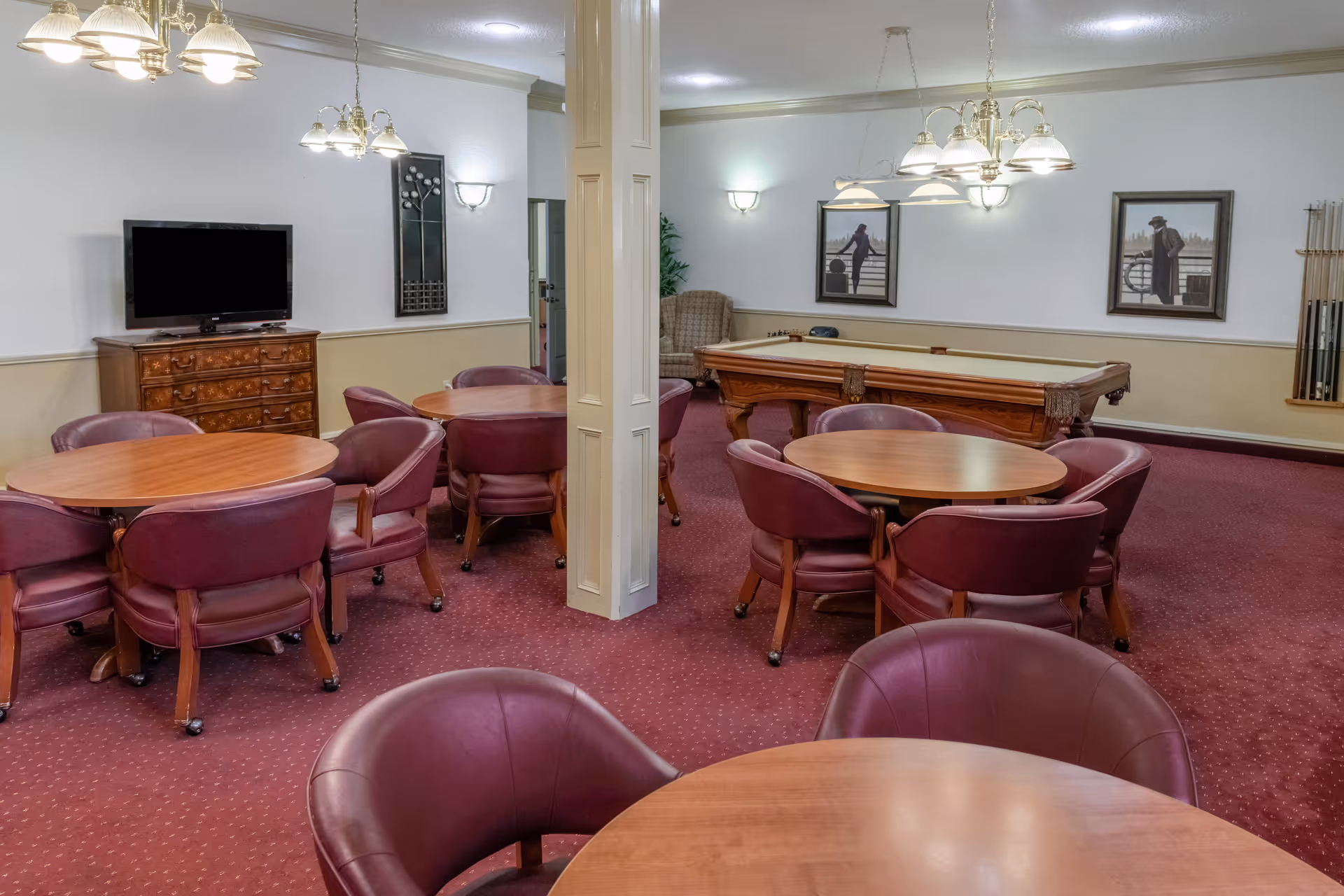 Carpeted community lounge with round wooden tables and burgundy chairs, a pool table, and a TV on a dresser.