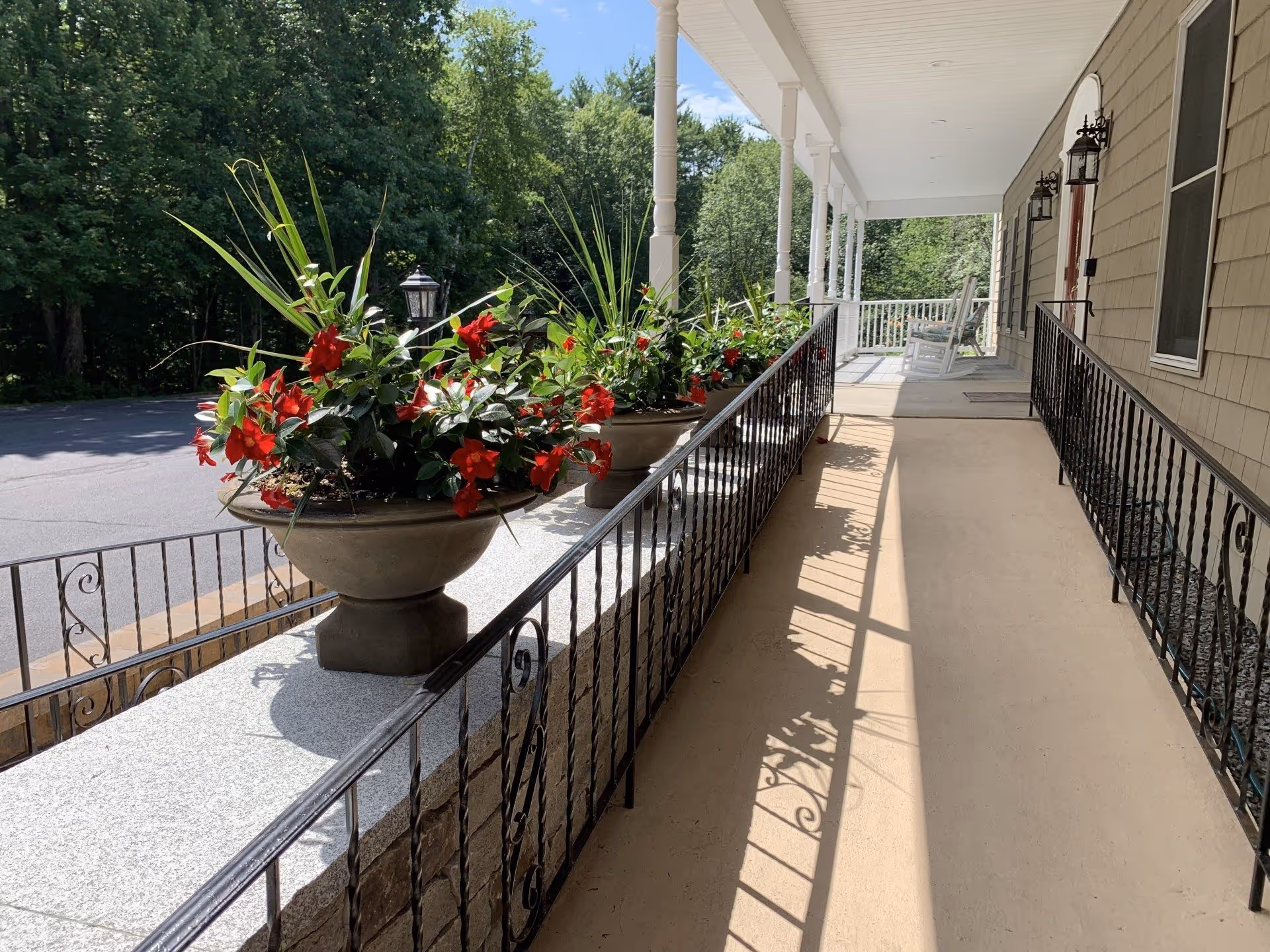 A sunny porch area with a beige floor and black wrought iron railings. Large planters with red flowers and green plants are placed along the railing. The porch has white columns and a white ceiling, with two rocking chairs visible in the distance. Trees and a paved area are visible beyond the porch.