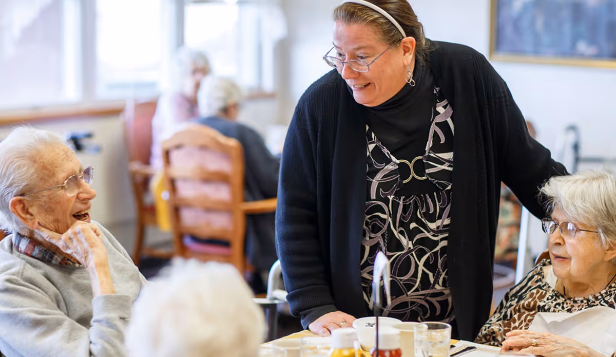 A staff member smiles and talks with elderly residents seated around a dining table in a communal dining area.