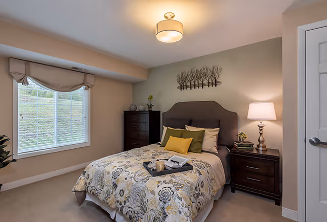 A cozy bedroom with a large window covered by a beige valance, a bed with a floral patterned comforter and multiple pillows, a dark wooden nightstand with a lamp and books, a matching tall dresser, and a decorative metal wall art above the bed.